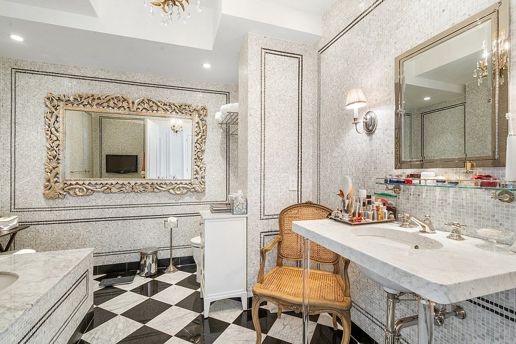This luxurious bathroom features a classic black-and-white checkered floor and intricate mosaic wall tiling. The space is anchored by a stone-topped vanity with a polished metal base and a large, ornate gold-framed mirror, complemented by a matching mirror on the adjacent wall. A vintage-style cane chair adds a touch of warmth to the sophisticated, high-contrast design.