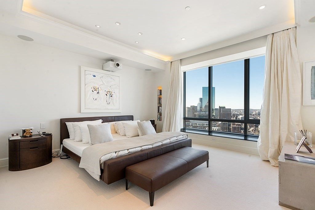 This sophisticated primary bedroom features a low-profile dark wood bed frame centered against a white wall adorned with a large piece of abstract art. A long, dark leather bench sits at the foot of the bed, while a wall of large windows provides an expansive, elevated view of the city skyline. The room is finished with neutral carpeting, recessed lighting, and elegant floor-length curtains that frame the urban vista.