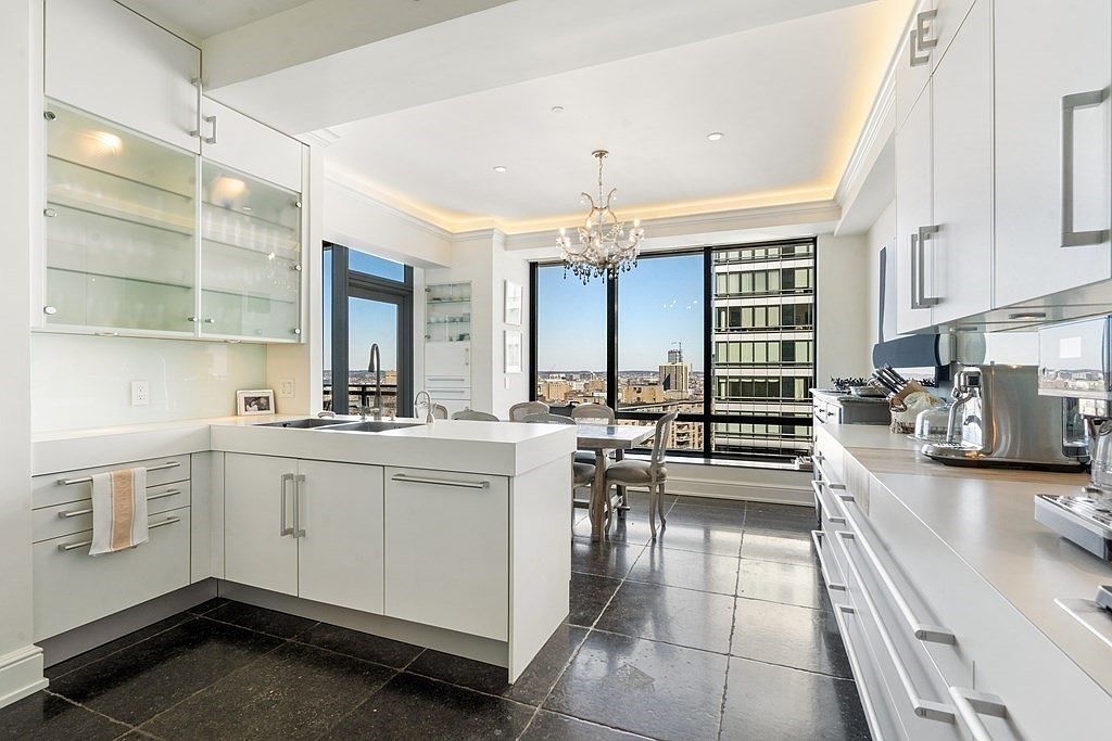 This bright, modern kitchen features sleek white cabinetry, a central island with a sink, and dark stone flooring that provides a sharp contrast. Large windows offer an expansive urban view, while a crystal chandelier hangs above the adjacent dining area, adding a touch of elegance. The space is clean and minimalist, emphasizing a high-end, contemporary aesthetic with ample natural light.