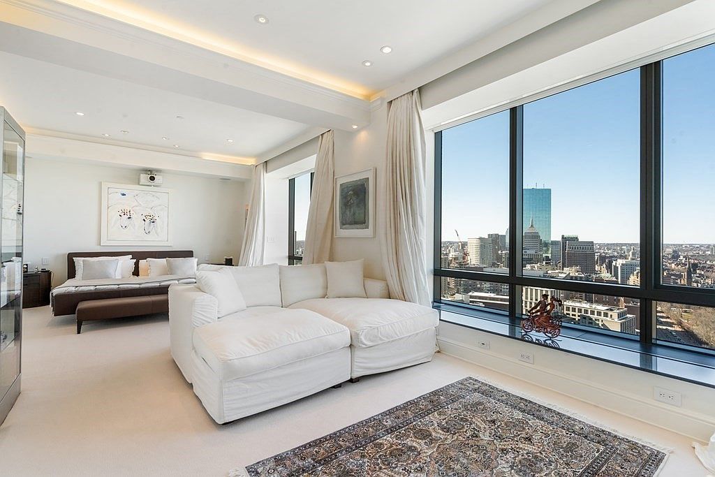This sophisticated primary bedroom suite features a seamless open-concept layout that integrates a plush white sectional sofa with a sleeping area. Large windows provide expansive, unobstructed views of the city skyline, bathing the room in natural light. The space is finished with neutral carpeting, elegant drapery, and a classic patterned area rug, creating a serene and luxurious urban retreat.