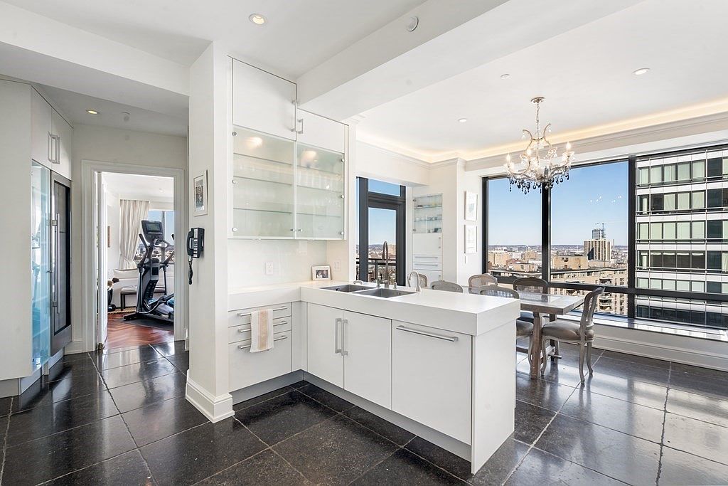 This modern, high-rise kitchen features sleek white cabinetry, a central island with a sink, and dark stone flooring that contrasts with the bright, minimalist aesthetic. Large windows provide an expansive urban view, while a crystal chandelier hangs over the adjacent dining area, adding a touch of elegance. The perspective captures the open-concept layout, looking from the kitchen toward the dining space and a glimpse of a home gym through the doorway.