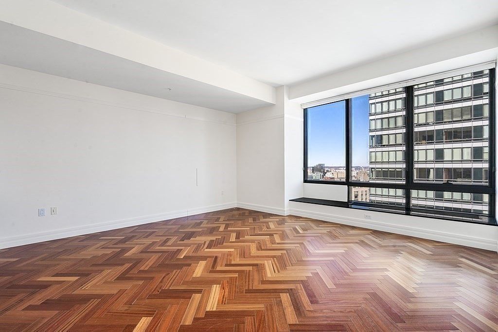 This bright, unfurnished room features rich, dark wood flooring laid in a classic herringbone pattern. A large window wall provides an expansive view of the surrounding city skyline, filling the space with natural light. The neutral white walls and minimalist design create a clean, versatile canvas for potential residents.