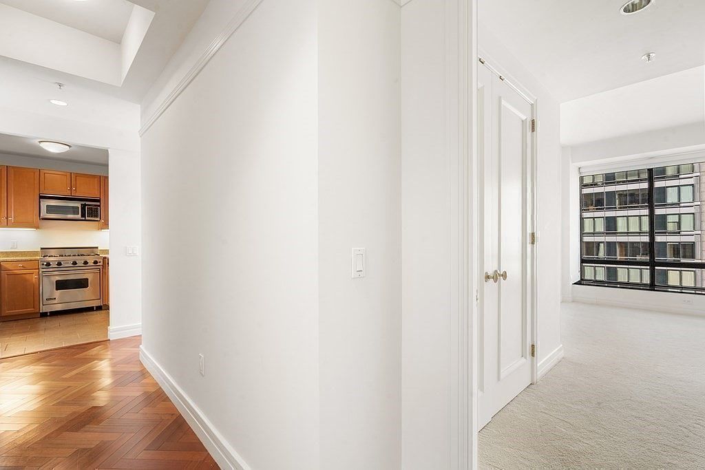 This interior view captures a transition space connecting a kitchen with wood herringbone flooring to a carpeted living area. A central white wall separates the two zones, featuring a closed door that leads to an adjacent room. The perspective looks through the hallway, highlighting the contrast between the warm wood tones of the kitchen and the bright, neutral palette of the living space.