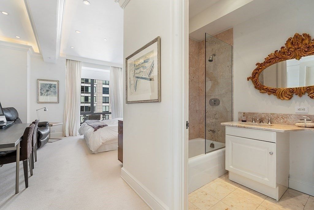 This interior view captures a transition space connecting a dining area, a bedroom, and a bathroom. To the left, a dining table is set against a wall with artwork, while a bedroom with large windows is visible in the background. To the right, a bathroom features a vanity with a decorative gold-toned mirror and a shower-tub combination with light-colored stone wall tiles.