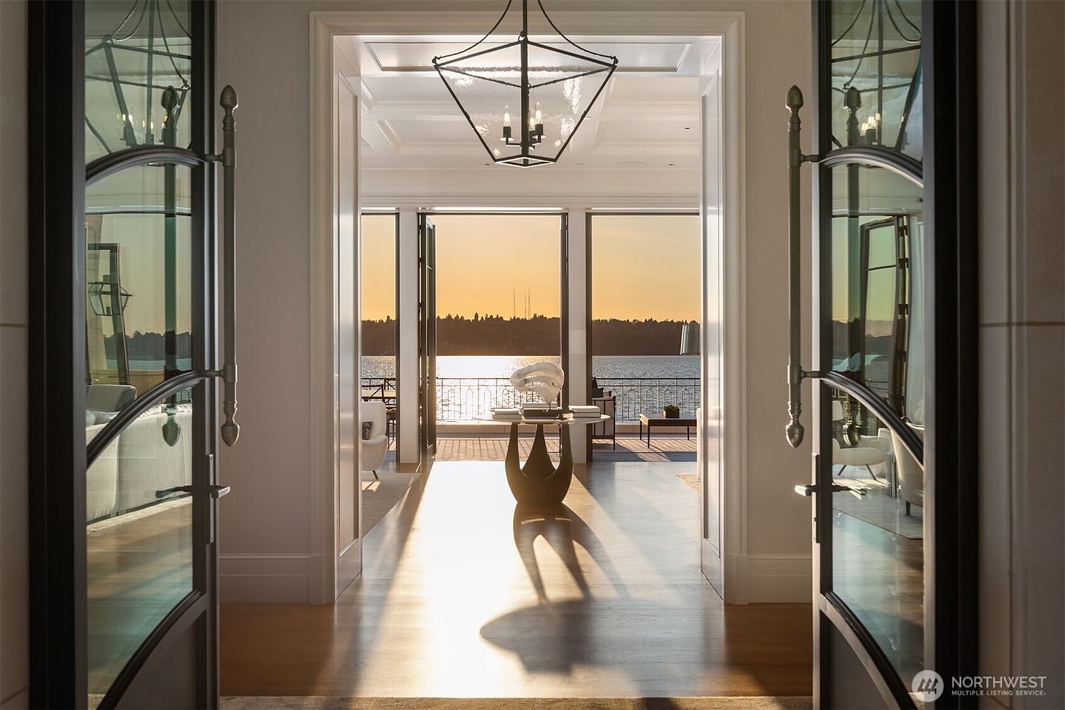 This interior shot showcases a grand hallway leading to a bright living space with waterfront views. The hallway features polished hardwood floors, elegant white trim, and a modern light fixture. Large glass doors on either side of the hallway reflect the interior, adding depth and a sense of luxury.