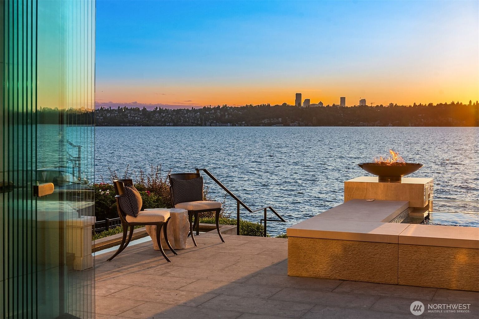 This image showcases a luxurious patio or deck area with stunning waterfront views. The space features modern outdoor furniture, including chairs and a built-in seating area with a fire pit. The overall impression is one of high-end relaxation and sophisticated outdoor living, perfect for enjoying the scenic surroundings.