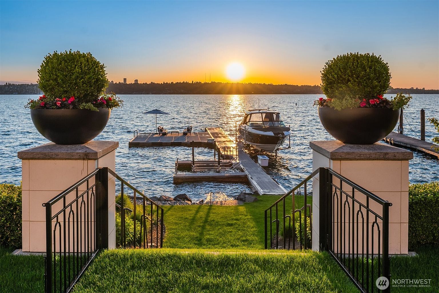 This stunning rear view showcases a waterfront property with a private dock and boat lift, set against a vibrant sunset. The manicured lawn leads to a staircase framed by decorative planters and wrought iron railings, offering a picturesque transition to the water. The scene exudes luxury and tranquility, highlighting the property's prime location and outdoor amenities.