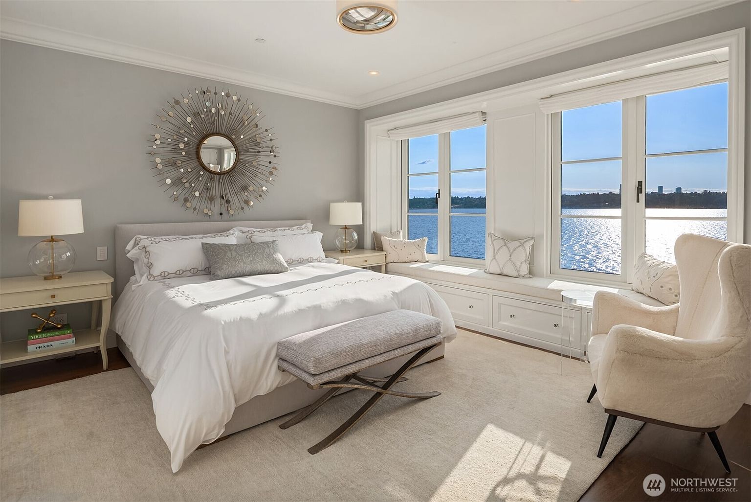 This is a bright and airy primary bedroom featuring a large bed with white linens, a decorative mirror above the headboard, and a window seat with a view of the water. The room is decorated in neutral tones with a light-colored rug and an accent chair, creating a serene and inviting atmosphere. The natural light floods the room, highlighting the clean lines and elegant design.