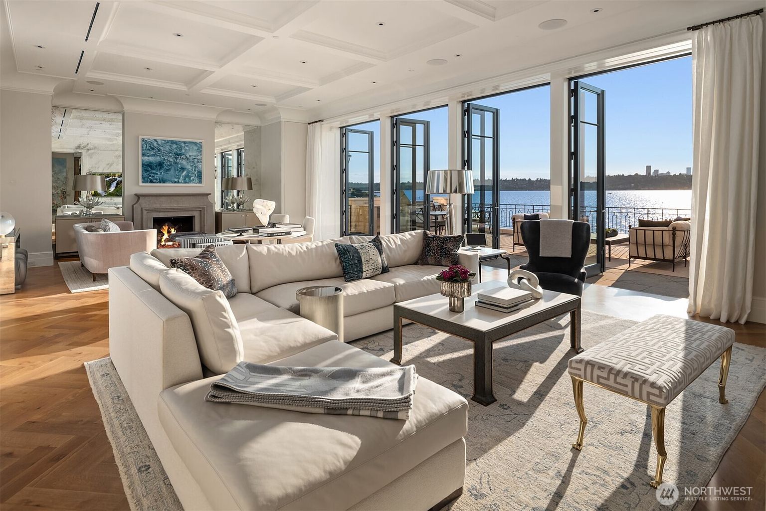 This is a bright and airy living room with a large sectional sofa, a coffee table, and a bench. The room features floor-to-ceiling windows that offer stunning water views and access to a patio. The overall style is luxurious and modern, creating a comfortable and inviting space.