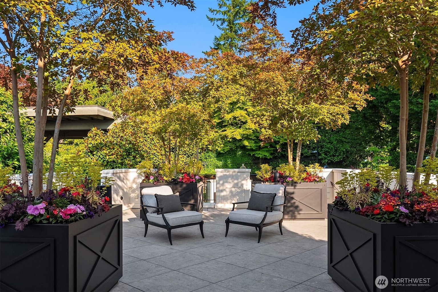 This image showcases a beautifully designed patio area, featuring two modern armchairs with gray cushions and throw pillows, placed on a tiled surface. Large, dark planters filled with colorful flowers and greenery frame the seating area, while trees with golden foliage provide a natural backdrop and a sense of privacy. The overall impression is one of a serene and stylish outdoor living space.
