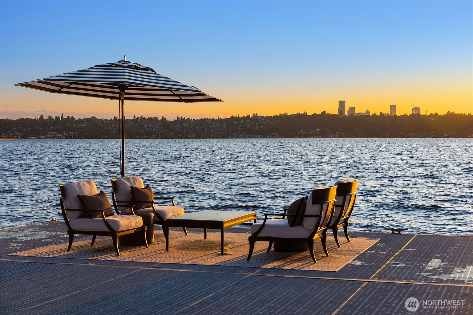 This image showcases a luxurious outdoor patio or deck area, furnished with comfortable seating arrangements under a striped umbrella, all set against a stunning waterfront backdrop. The scene is bathed in the warm hues of a sunset, with a cityscape visible in the distance, creating an inviting and serene atmosphere perfect for relaxation and entertainment. The perspective is wide, capturing the vastness of the water and the skyline.