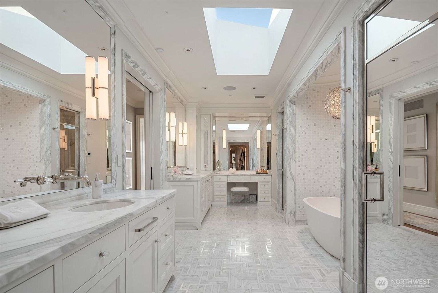 This is a luxurious primary bathroom featuring a double vanity with marble countertops, mosaic tile walls, and a herringbone-patterned marble floor. The bathroom is well-lit with a skylight and elegant sconces, and includes a freestanding bathtub and separate shower area. The overall impression is one of opulence and tranquility.