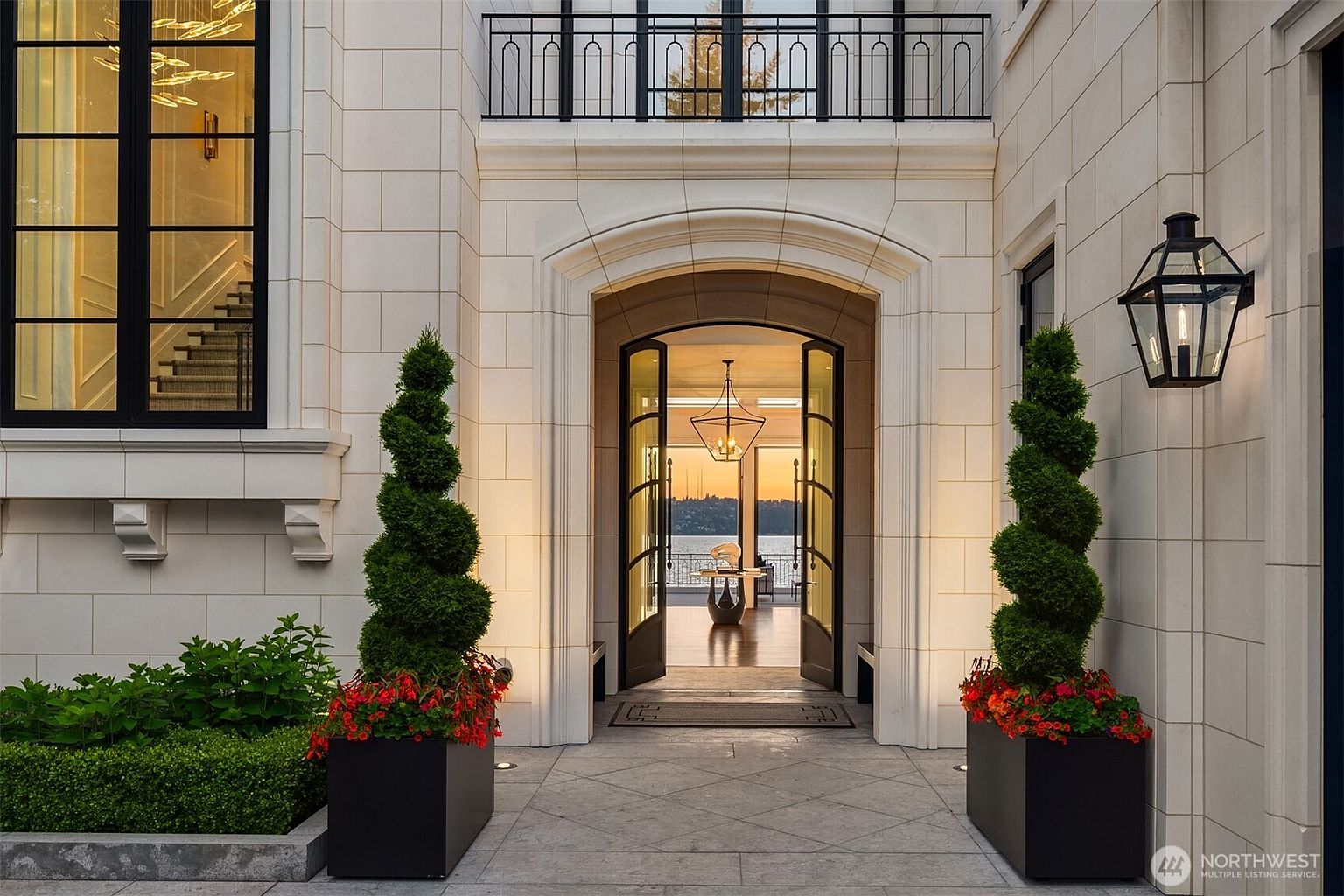The image showcases the grand entryway of a luxurious home, featuring an arched doorway with double doors that open to reveal a glimpse of the interior and a waterfront view. Flanking the entrance are meticulously manicured spiral topiaries in square planters, adding a touch of elegance and symmetry. The exterior is clad in light-colored stone, complemented by black-framed windows and a balcony above the entrance.