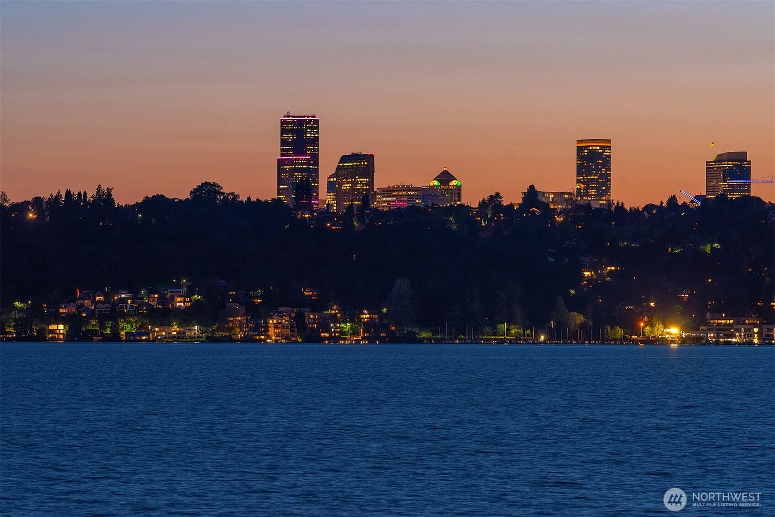 This image showcases a stunning cityscape at dusk, featuring illuminated buildings against a colorful sky. The scene is viewed from across a body of water, with waterfront homes visible along the shoreline. The overall impression is one of urban sophistication and serene waterfront living.