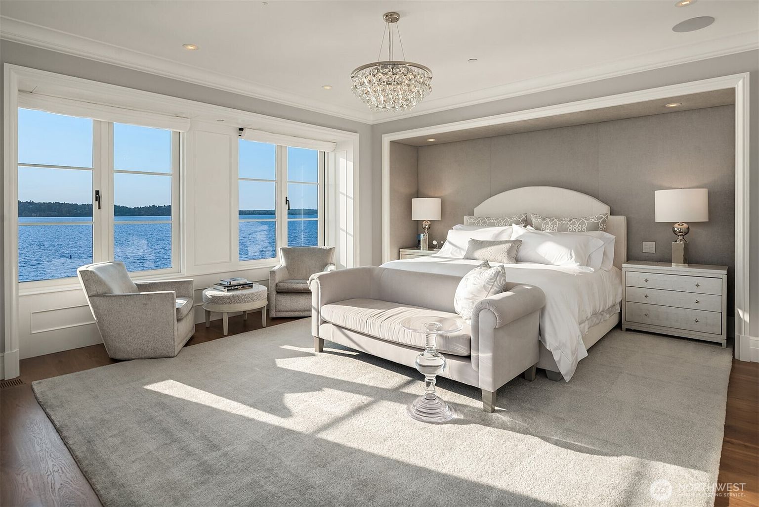 This is a luxurious primary bedroom featuring a large bed with white linens, a sitting area with two chairs and a small table by the windows, and a crystal chandelier. The room is decorated in neutral tones with gray walls and a light gray rug, creating a serene and elegant atmosphere. Large windows offer a view of the water, enhancing the room's appeal.