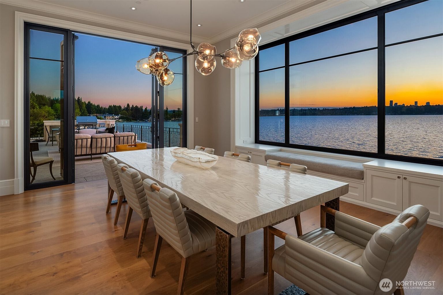 This is an interior shot of a dining room featuring a large, light-colored wooden table with modern chairs. A contemporary chandelier hangs above the table, and a large window offers a stunning view of the water and city skyline at sunset. The room opens onto a patio with outdoor seating, creating a seamless indoor-outdoor living experience.