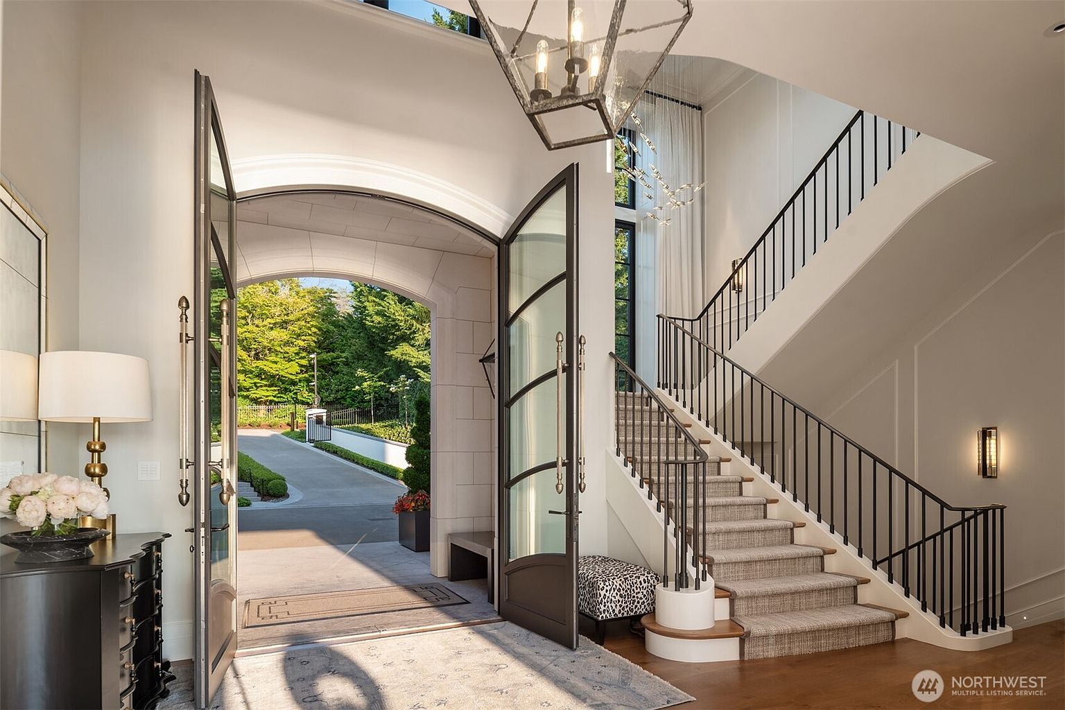 This interior shot showcases a grand entryway with an open front door leading to a driveway lined with greenery. The foyer features a striking staircase with black metal railings, complemented by a modern light fixture and a patterned ottoman. The overall impression is one of luxury and sophisticated design.