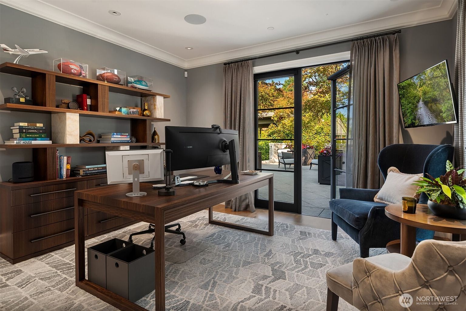 This is a well-appointed home office featuring a large wooden desk with modern computer equipment, custom built-in shelving displaying decorative items and books, and comfortable seating. The room is bathed in natural light from the adjacent patio, creating a serene and productive workspace. The neutral color palette and sophisticated furnishings contribute to a professional and inviting atmosphere.