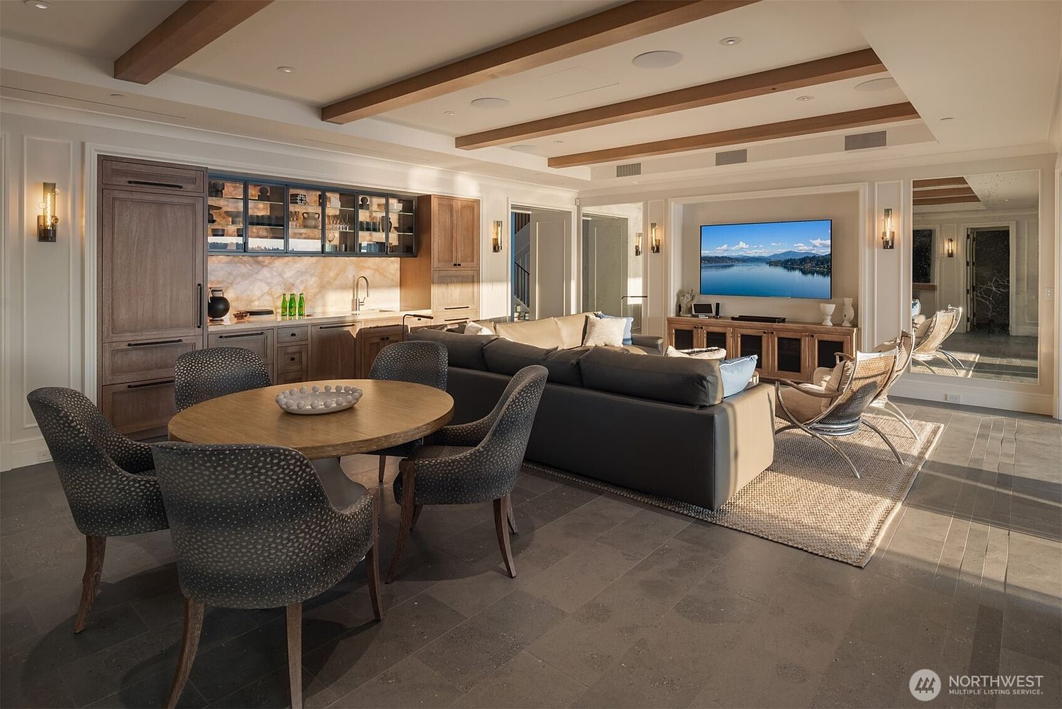This interior shot showcases a stylish living room with a modern aesthetic. The room features a comfortable sectional sofa, a round dining table with chairs, a built-in bar area, and a large television mounted above a wooden console. The space is well-lit with recessed lighting and sconces, and the overall impression is one of sophisticated comfort and functionality.