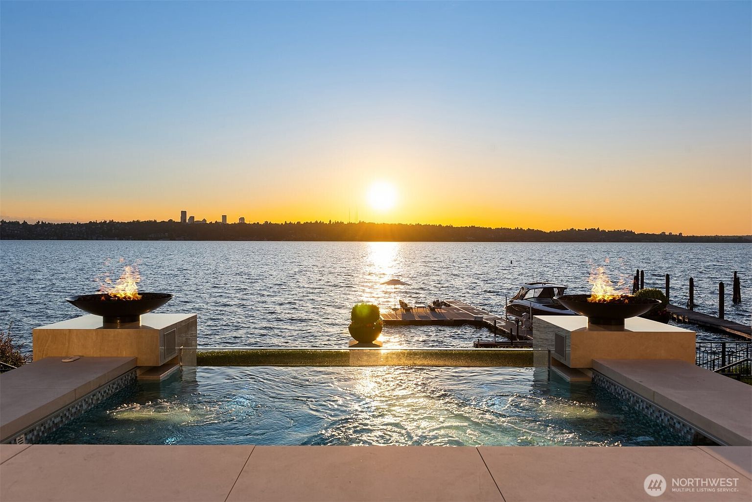 This image showcases a luxurious pool and spa area overlooking a serene body of water at sunset. The pool features modern design elements, including fire bowls on either side and a seamless edge. A boat dock is visible in the background, adding to the property's appeal and suggesting waterfront access.