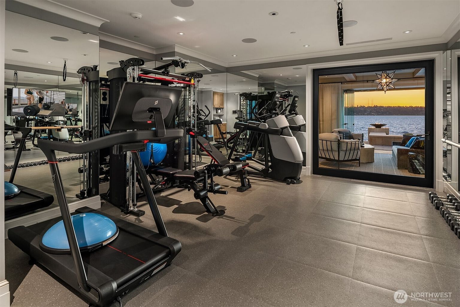 This is a well-equipped home gym featuring a treadmill, weight machines, and free weights. The room has mirrored walls, providing a sense of spaciousness, and a large sliding glass door offers a view of the water. The flooring is a dark, durable material, and the overall impression is one of a high-end, functional workout space.