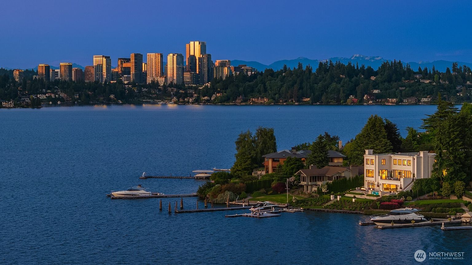 This aerial view showcases a luxurious waterfront property with a modern white house, private docks, and several boats. The property is nestled on a lush green peninsula, with a stunning cityscape and mountain range visible in the background. The image conveys a sense of exclusivity and high-end living.