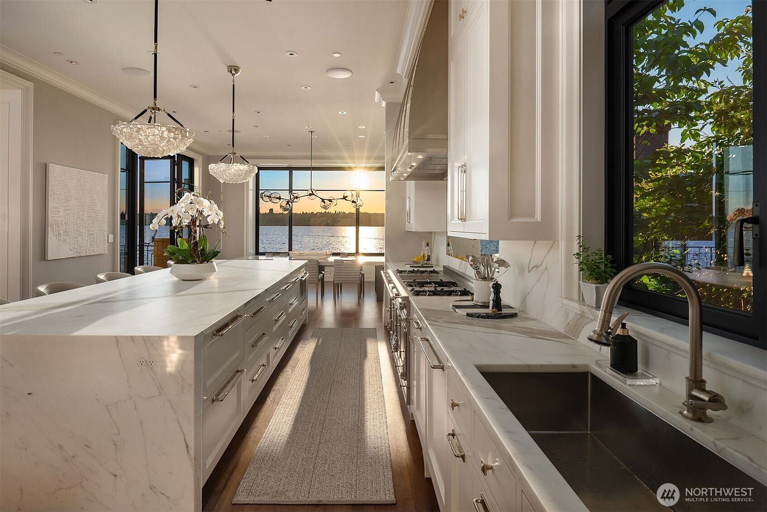 This is a bright and luxurious kitchen featuring white marble countertops and cabinetry, complemented by stainless steel appliances and fixtures. Large windows offer a stunning view of the water, enhancing the kitchen's appeal. The space is illuminated by elegant pendant lighting, creating a sophisticated and inviting atmosphere.