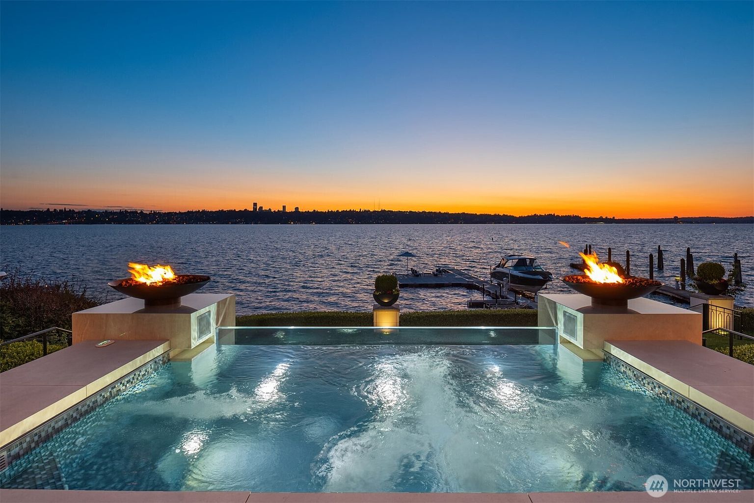 This image showcases a luxurious pool and spa area with a stunning waterfront view at sunset. The pool features clear glass edges, and fire bowls add a touch of elegance and warmth. A boat dock is visible in the background, enhancing the property's appeal for waterfront living.