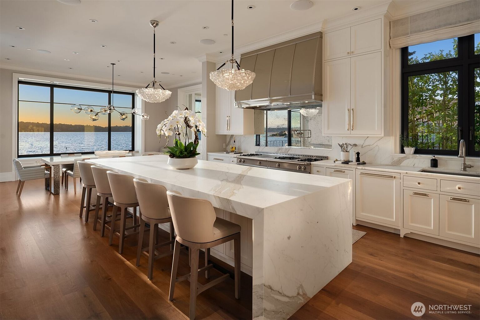 This is a bright and spacious kitchen featuring a large marble island with seating, perfect for entertaining. The kitchen boasts white cabinetry, stainless steel appliances, and a large window offering a stunning view of the water. The open layout seamlessly connects the kitchen to the dining area, creating a cohesive and inviting space.