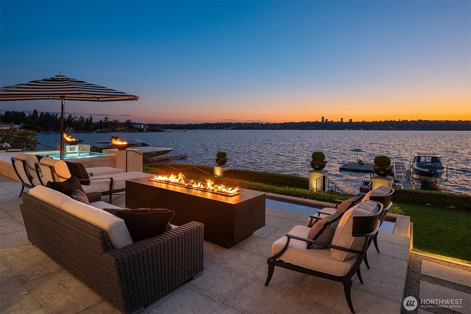 This image showcases a luxurious outdoor patio area at dusk, featuring comfortable seating around a modern fire pit. The patio overlooks a serene lake with a cityscape visible in the distance, creating a tranquil and upscale ambiance. Additional amenities include a pool and a covered lounge area, enhancing the property's appeal for relaxation and entertainment.