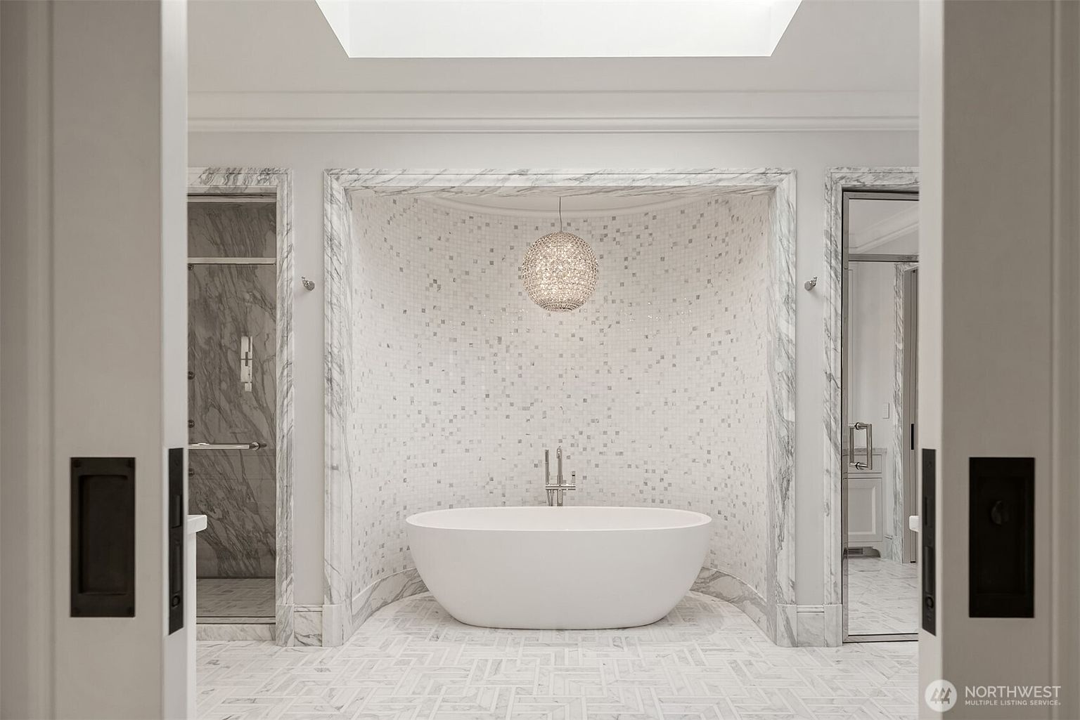 This is a luxurious primary bathroom featuring a freestanding white bathtub centered within a marble-tiled alcove, illuminated by a decorative pendant light. The bathroom showcases a modern design with clean lines, marble accents, and a spacious layout, creating a spa-like atmosphere. The view is from the doorway, looking into the bathroom.