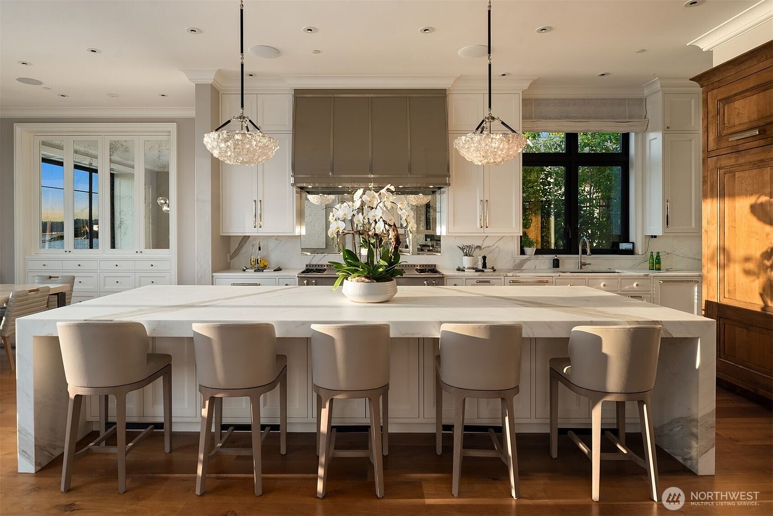 This is a bright and spacious kitchen featuring a large marble island with seating for five. The kitchen is equipped with stainless steel appliances, white cabinetry, and elegant pendant lighting. A large window provides natural light and a view of the outdoors, while a mirrored backsplash adds depth and visual interest.