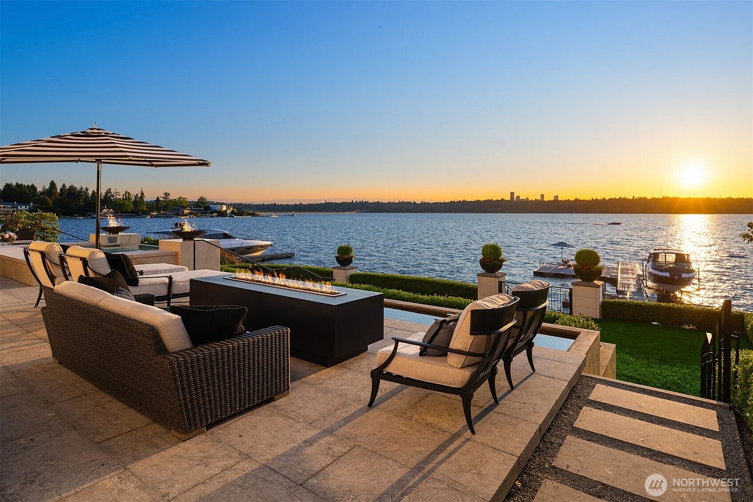 This image showcases a luxurious outdoor patio area with comfortable seating arrangements, including a wicker sofa and elegant chairs, centered around a modern fire pit. The patio overlooks a serene lake at sunset, with boats docked nearby and a cityscape visible in the distance. The scene exudes a sense of upscale relaxation and waterfront living.