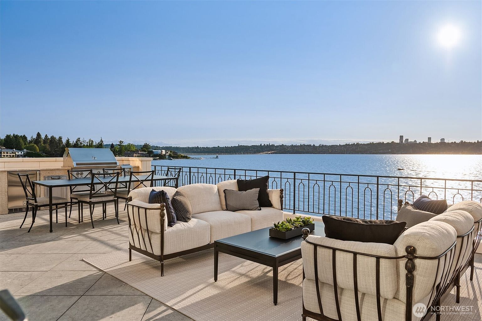 This image showcases a luxurious outdoor patio or balcony area with stunning waterfront views. The space is furnished with comfortable seating, including a sofa and armchairs, arranged around a coffee table. A built-in grill and dining set suggest an area perfect for entertaining, while the railing provides safety without obstructing the scenic vista.