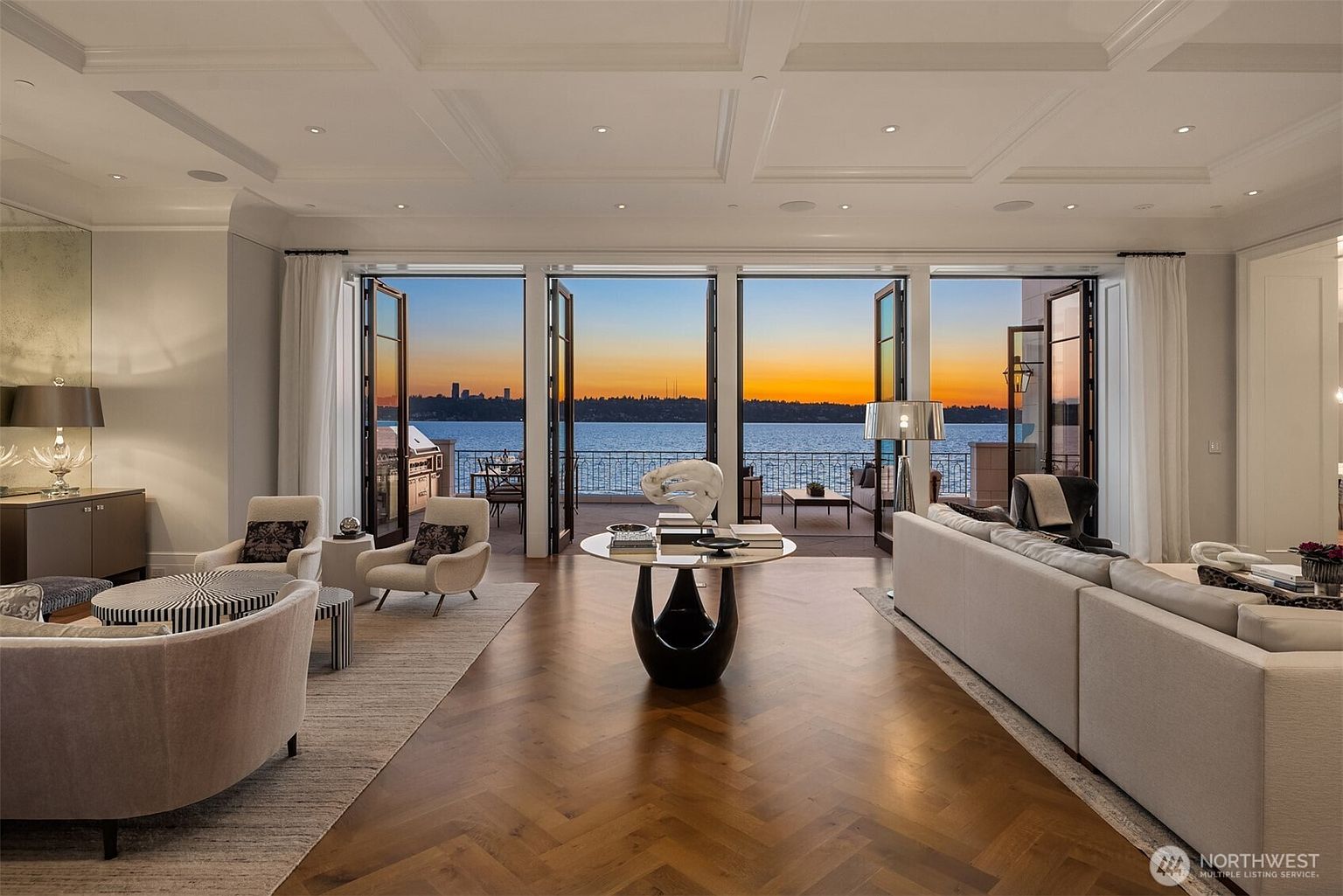 This is an interior shot of a luxurious living room with an open floor plan that leads to an outdoor balcony overlooking a body of water at sunset. The room features high ceilings with detailed molding, hardwood floors in a herringbone pattern, and elegant furniture including a large sofa and armchairs. The open doors to the balcony provide a seamless transition between indoor and outdoor living spaces.