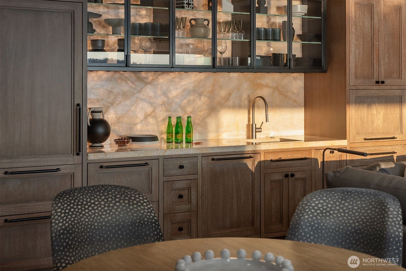 This interior shot showcases a modern kitchen area with wooden cabinetry and a light marble backsplash. Glass-front cabinets display dishware, while a sleek faucet and sink are set into the countertop. The scene is warmly lit, highlighting the textures and finishes, and a dining table with chairs is in the foreground.
