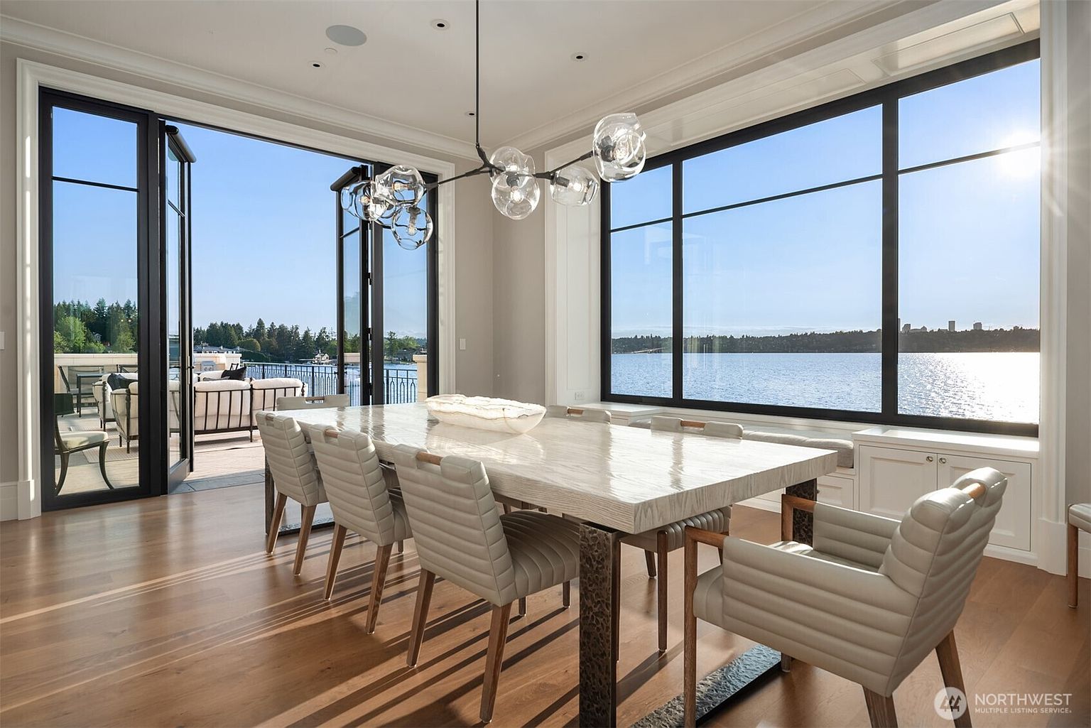 This is a bright and airy dining room featuring a large, modern dining table with seating for eight. The room boasts a large window with a view of the water and an open doorway leading to an outdoor patio. The hardwood floors and contemporary lighting fixture add to the room's elegant and inviting atmosphere.