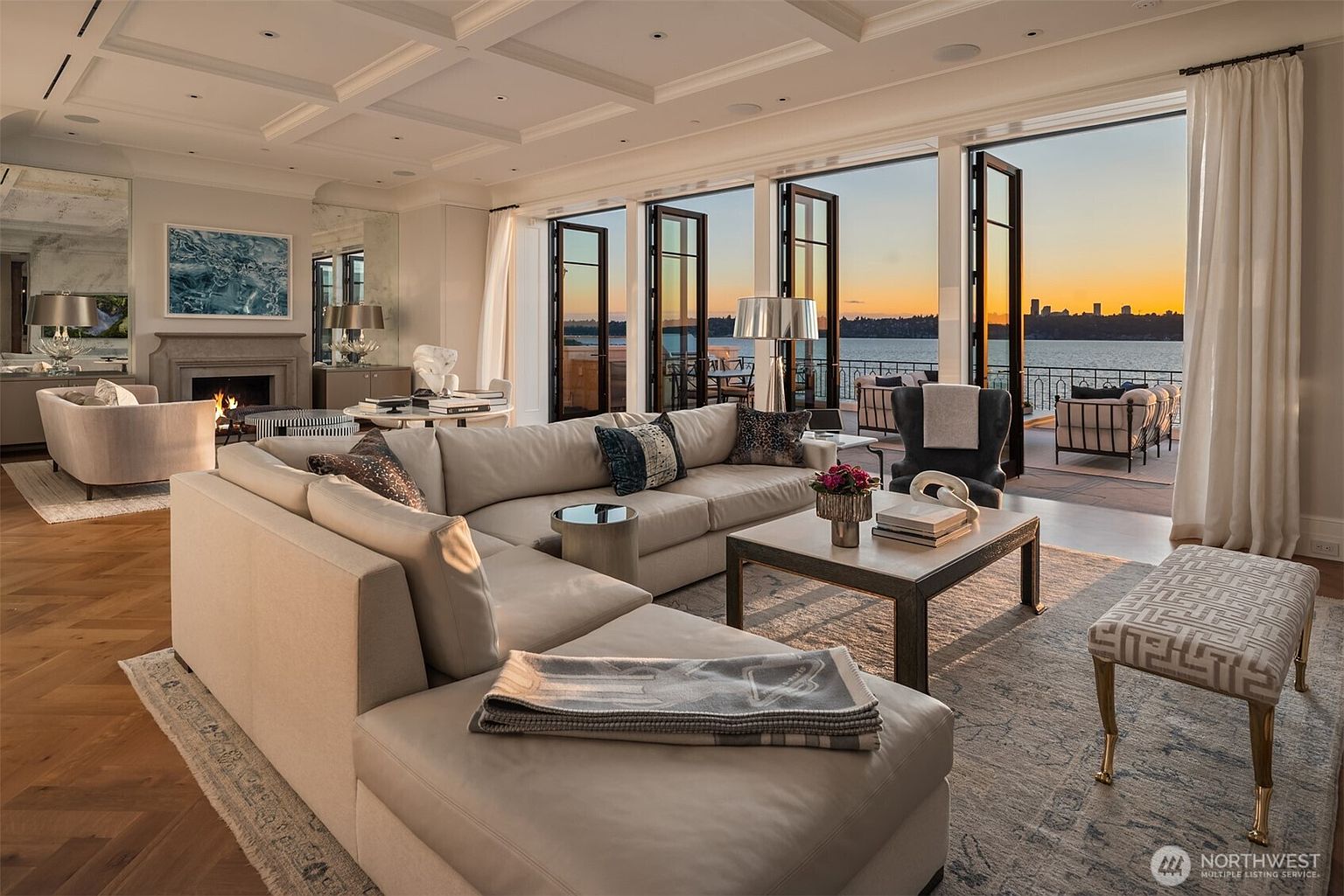 This is a luxurious living room featuring a large sectional sofa, a coffee table with decorative items, and a view of the water through large windows. The room is decorated in neutral tones with pops of color from the pillows and flowers. The overall impression is one of elegance and comfort, perfect for relaxing and entertaining.