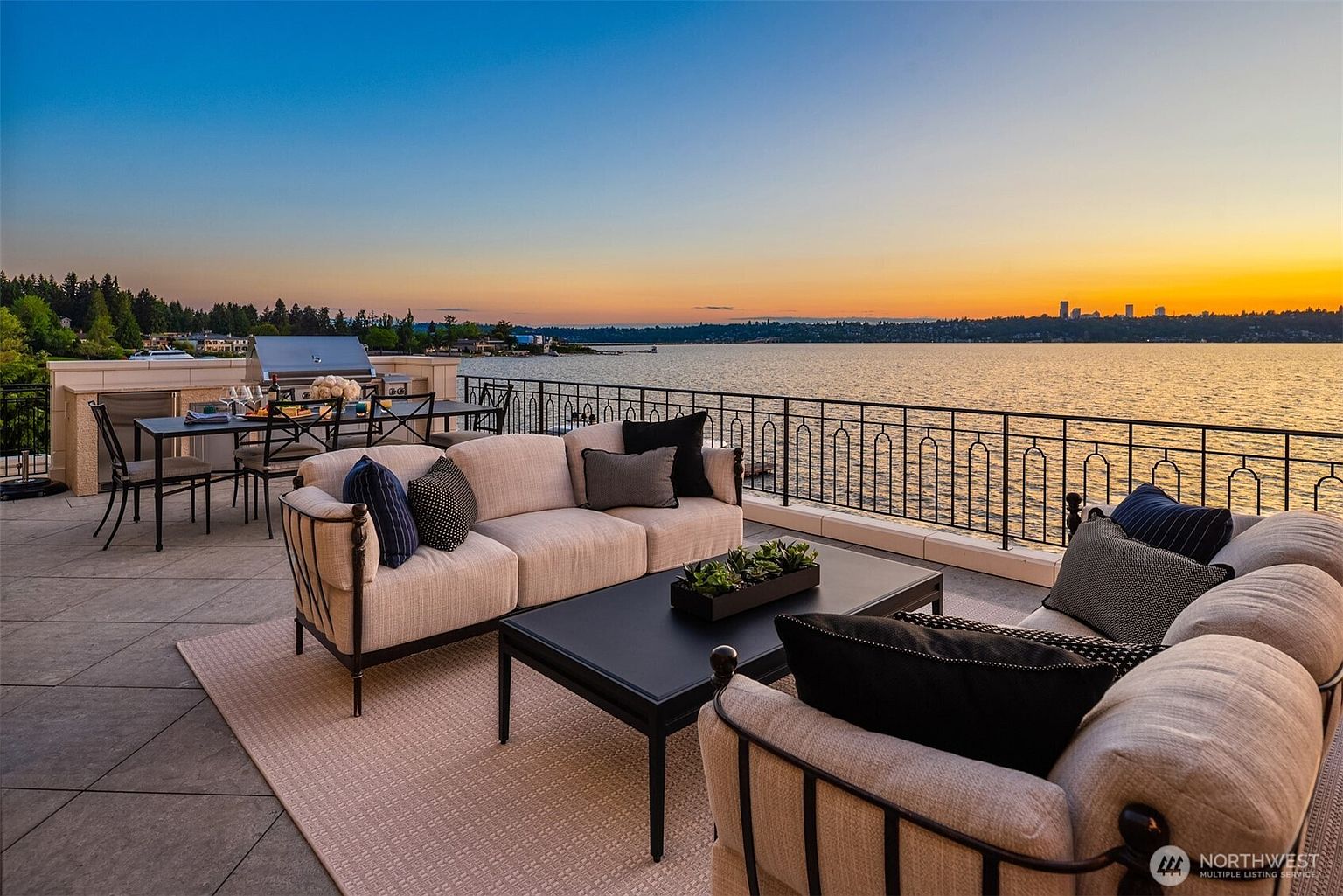 This image showcases a luxurious outdoor patio or deck area, featuring comfortable seating arrangements with plush sofas and chairs, complemented by a sleek coffee table. The space is elegantly furnished and overlooks a stunning waterfront view at sunset, with a glimpse of a city skyline in the distance. A dining area with a grill is also visible, suggesting an ideal space for outdoor entertaining.