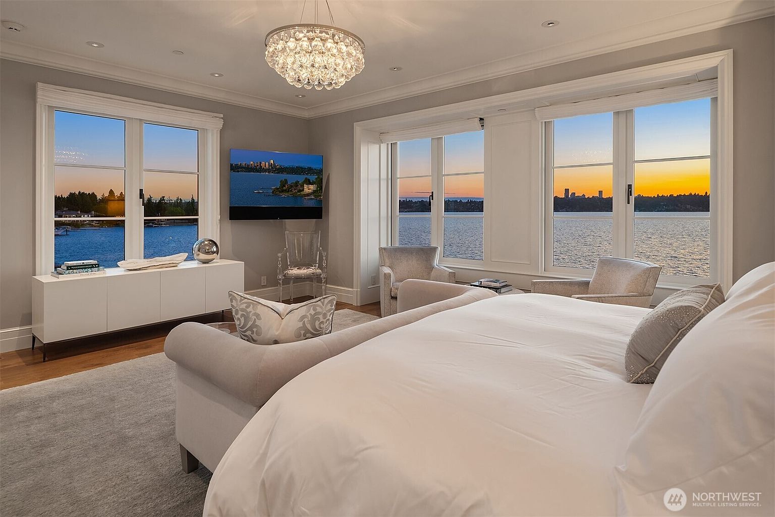 This is a luxurious primary bedroom featuring large windows with stunning water views and a glimpse of the city skyline at sunset. The room is elegantly furnished with a plush bed, comfortable seating, and a modern chandelier, creating a serene and upscale ambiance. A sleek white console table and a wall-mounted television add to the room's functionality and style.
