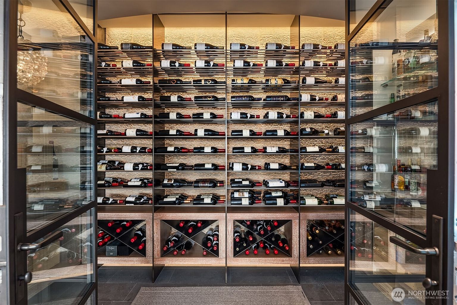 This image showcases a well-stocked wine cellar, featuring numerous bottles displayed on horizontal racks and in diamond-patterned storage. The cellar is enclosed by glass doors with dark metal frames, providing a clear view of the collection. The lighting highlights the textured backdrop, creating an inviting and sophisticated atmosphere, perfect for wine enthusiasts.
