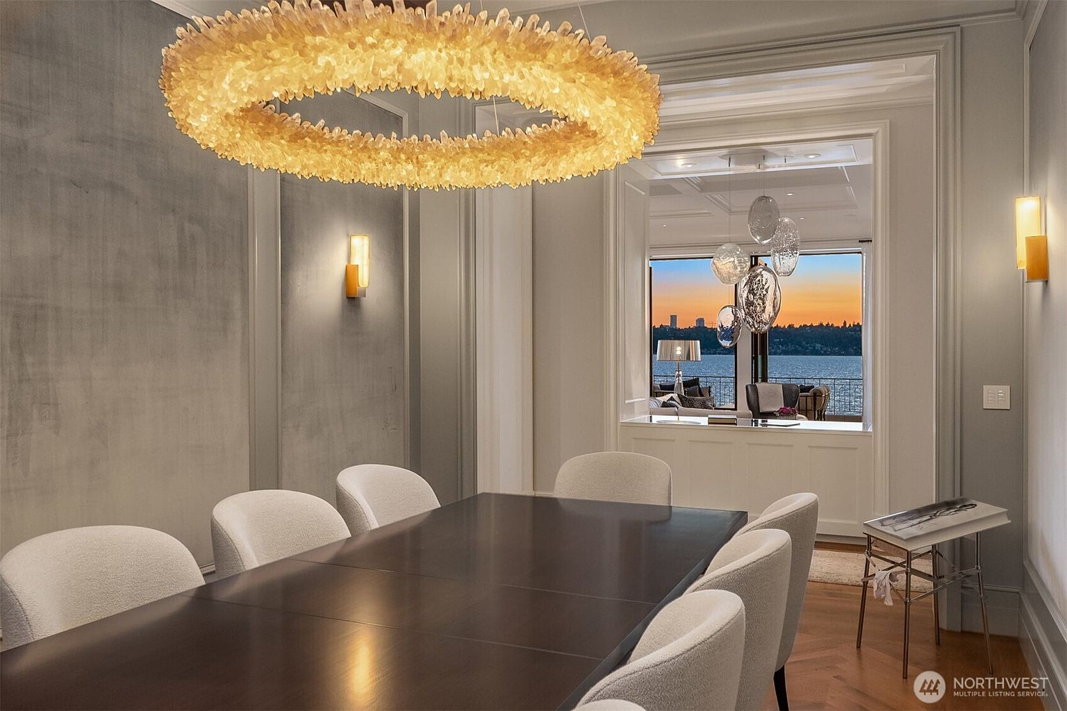 This is an interior shot of a dining room featuring a large, dark wood table surrounded by white upholstered chairs. A striking, circular chandelier hangs above the table, providing warm lighting. In the background, a window offers a view of a body of water and a cityscape at sunset, adding a touch of elegance and serenity to the space.