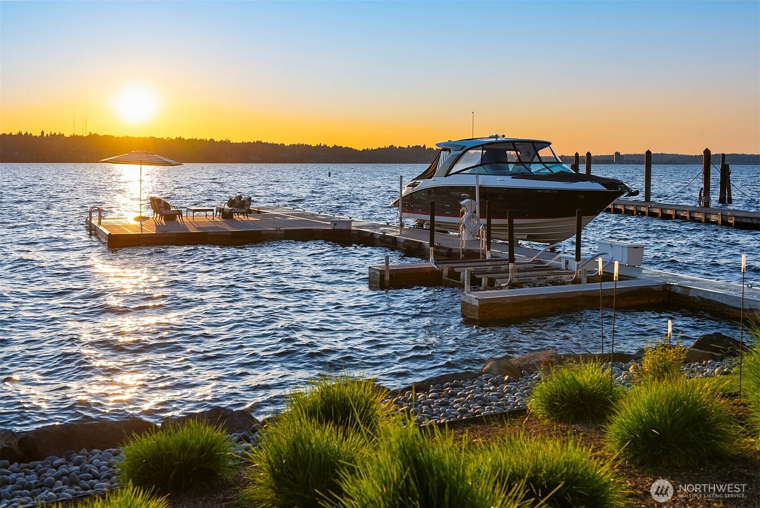 This image showcases a luxurious waterfront property featuring a private dock with a boat lift and a modern boat. The dock includes a seating area with an umbrella, perfect for enjoying the sunset over the calm water. Lush greenery in the foreground adds to the serene and upscale ambiance.