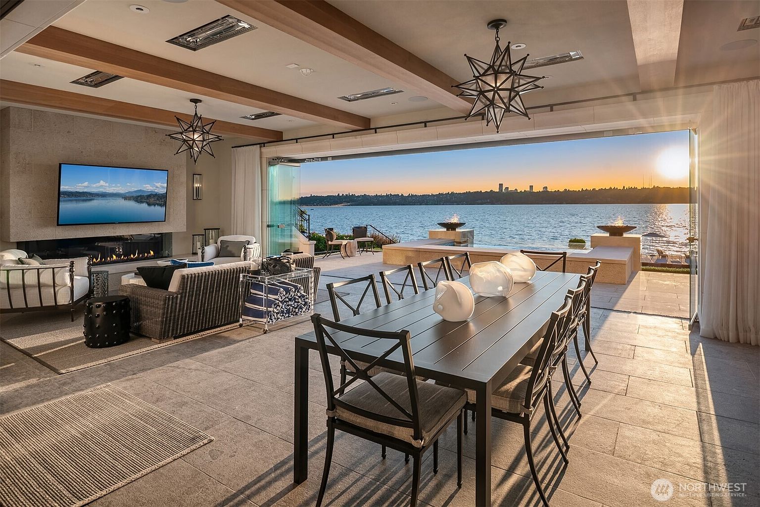This image showcases a luxurious living room that seamlessly blends indoor and outdoor spaces. The room features a stone fireplace with a large television, comfortable seating, and a dining area with a long table and modern light fixtures. Large sliding glass doors open to a patio with fire pits and a stunning waterfront view, creating an inviting and sophisticated atmosphere.