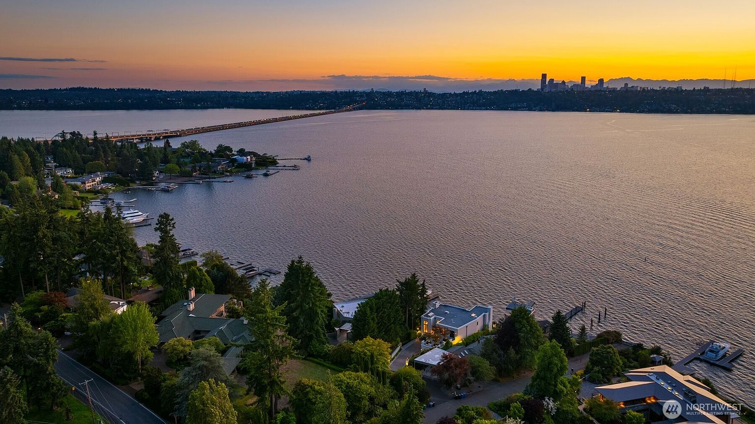 This aerial view showcases a stunning waterfront property at sunset. The home is surrounded by lush greenery and mature trees, with a private dock extending into the calm water. In the distance, a bridge connects to the city skyline, creating a picturesque backdrop that highlights the property's prime location and luxurious setting.