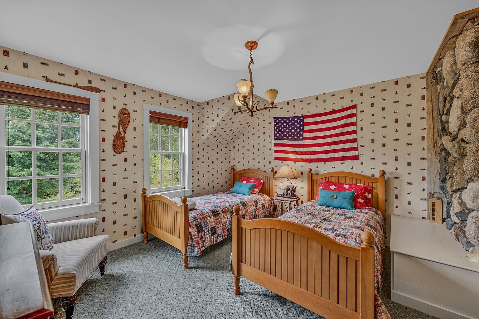 This cozy guest bedroom features two twin beds with patterned quilts and a rustic, lodge-inspired aesthetic. The room is accented by wallpaper with small motifs, a unique antler-style chandelier, and a prominent stone-clad chimney breast on the right wall. Two windows provide natural light, while a comfortable armchair sits in the corner, creating a warm and inviting atmosphere.