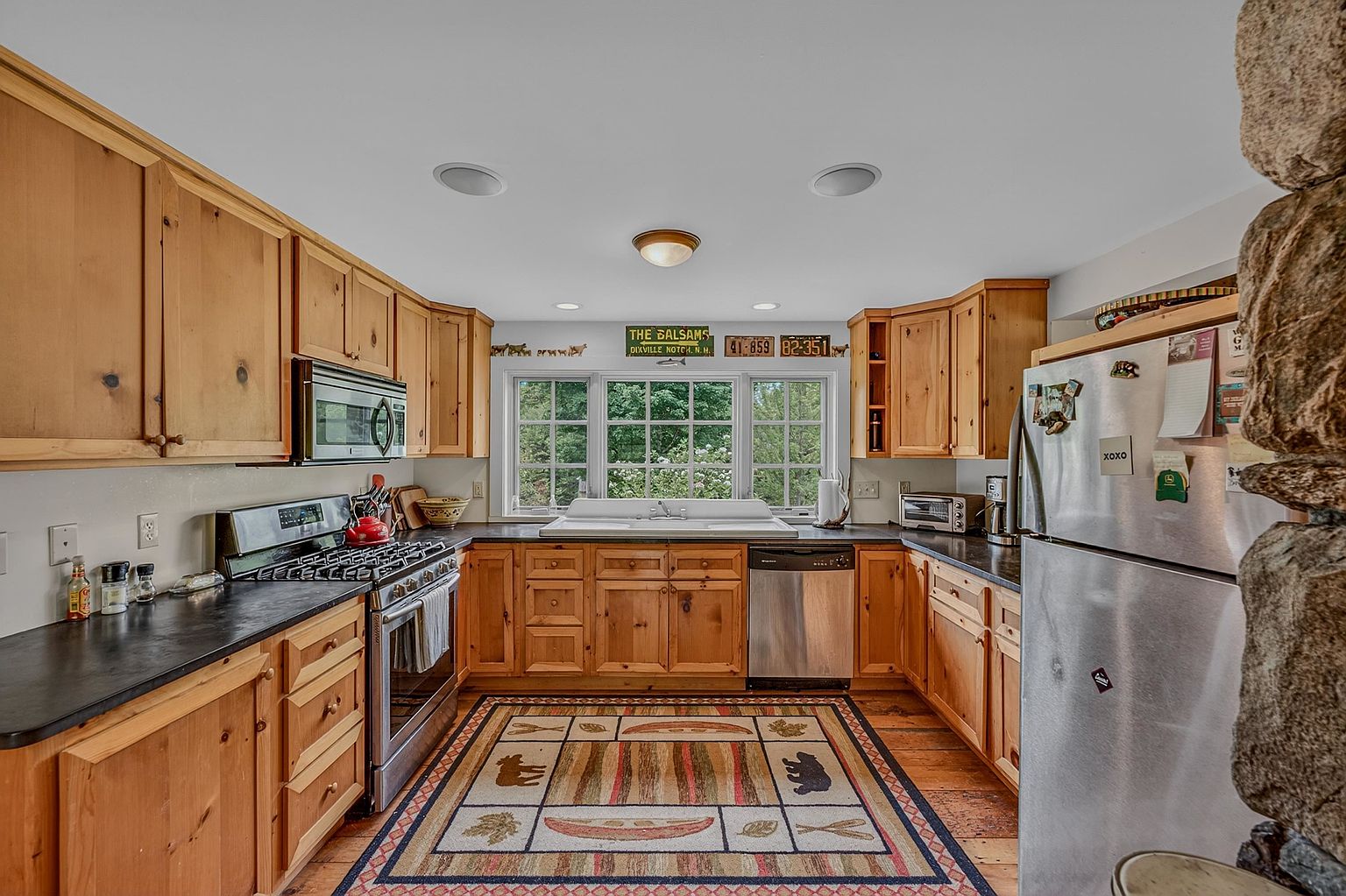 This rustic-style kitchen features warm light wood cabinetry paired with dark stone countertops and a suite of stainless steel appliances. A large, multi-pane window above the sink provides natural light, while a decorative area rug with wildlife motifs adds character to the space. To the right, a prominent stone-clad chimney breast adds a textured, organic element to the room's cozy, cabin-inspired aesthetic.