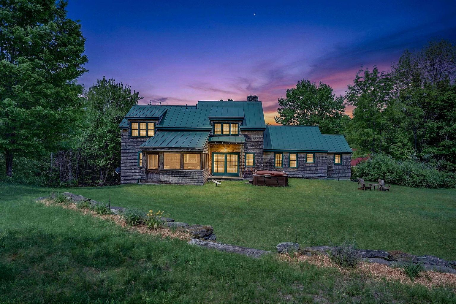 This rear view of a shingle-style home showcases a classic aesthetic with a prominent green metal roof and a sprawling, well-manicured lawn. The house features a mix of multi-pane windows, a screened-in porch area, and a hot tub situated on the grass near the structure. A rustic stone retaining wall defines the foreground, while the vibrant sunset sky provides a dramatic backdrop for the property.