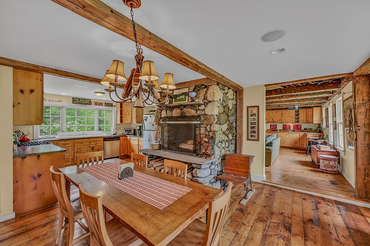 This rustic dining area features a large wooden table centered beneath a multi-armed chandelier, set against a prominent stone-clad chimney breast that serves as a focal point. The space flows openly into a kitchen with light wood cabinetry and a separate living area visible through a wide doorway, all tied together by warm wood flooring and exposed ceiling beams. The overall aesthetic is cozy and traditional, emphasizing natural textures and a welcoming, open-concept layout.