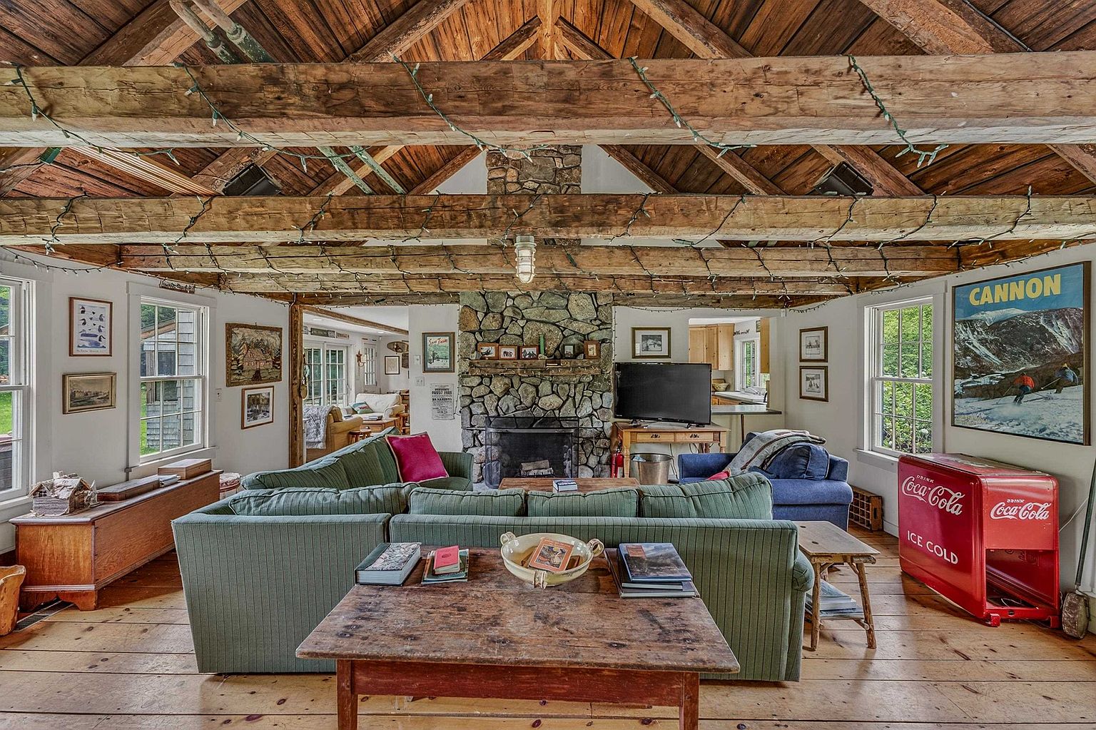 This rustic living room features a vaulted ceiling with exposed timber beams and string lights, creating a cozy, cabin-like atmosphere. A prominent stone-clad chimney breast serves as the focal point, complemented by a large green sectional sofa and a vintage-style red Coca-Cola cooler. The space is finished with warm wood flooring and a collection of framed wall art, offering a comfortable and nostalgic living environment.